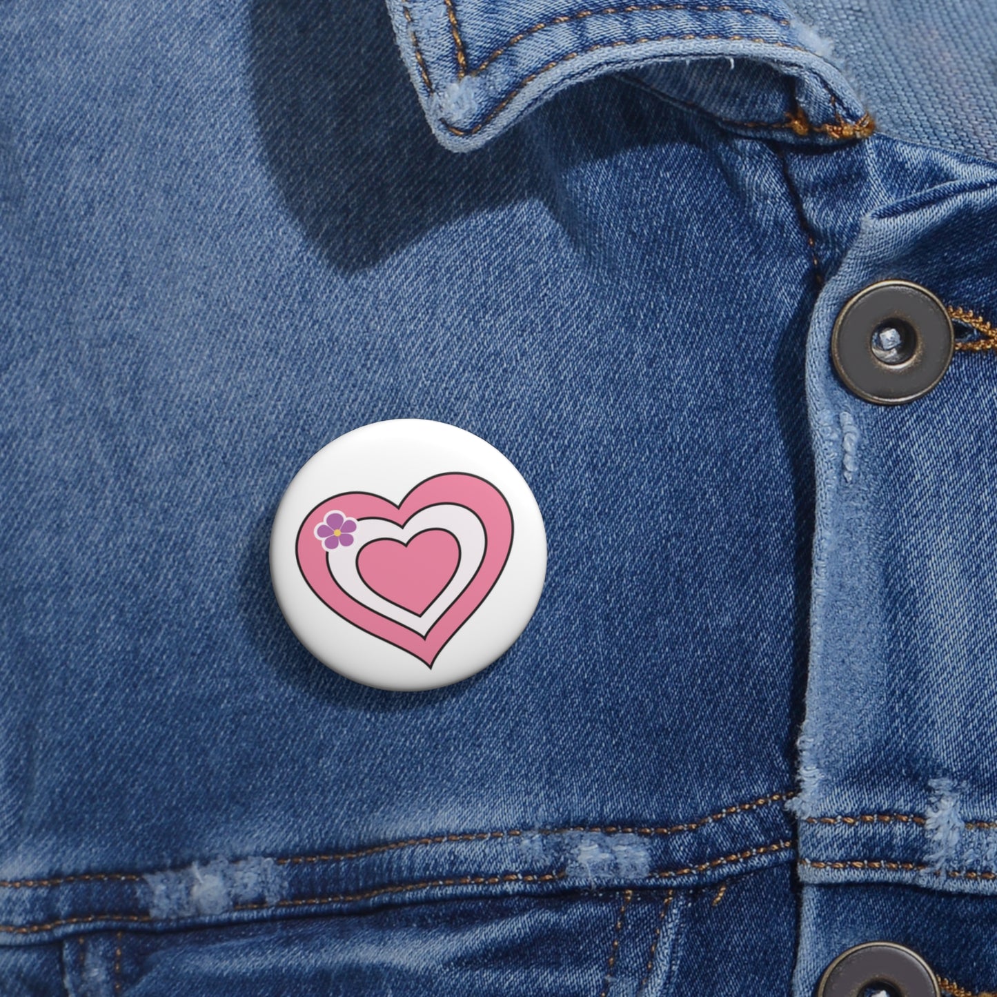 Close-up of a white circular pride button featuring a nested heart design inspired by the Sapphic pride flag in pink and white, attached to a blue denim jacket at the right chest with the button clearly visible.