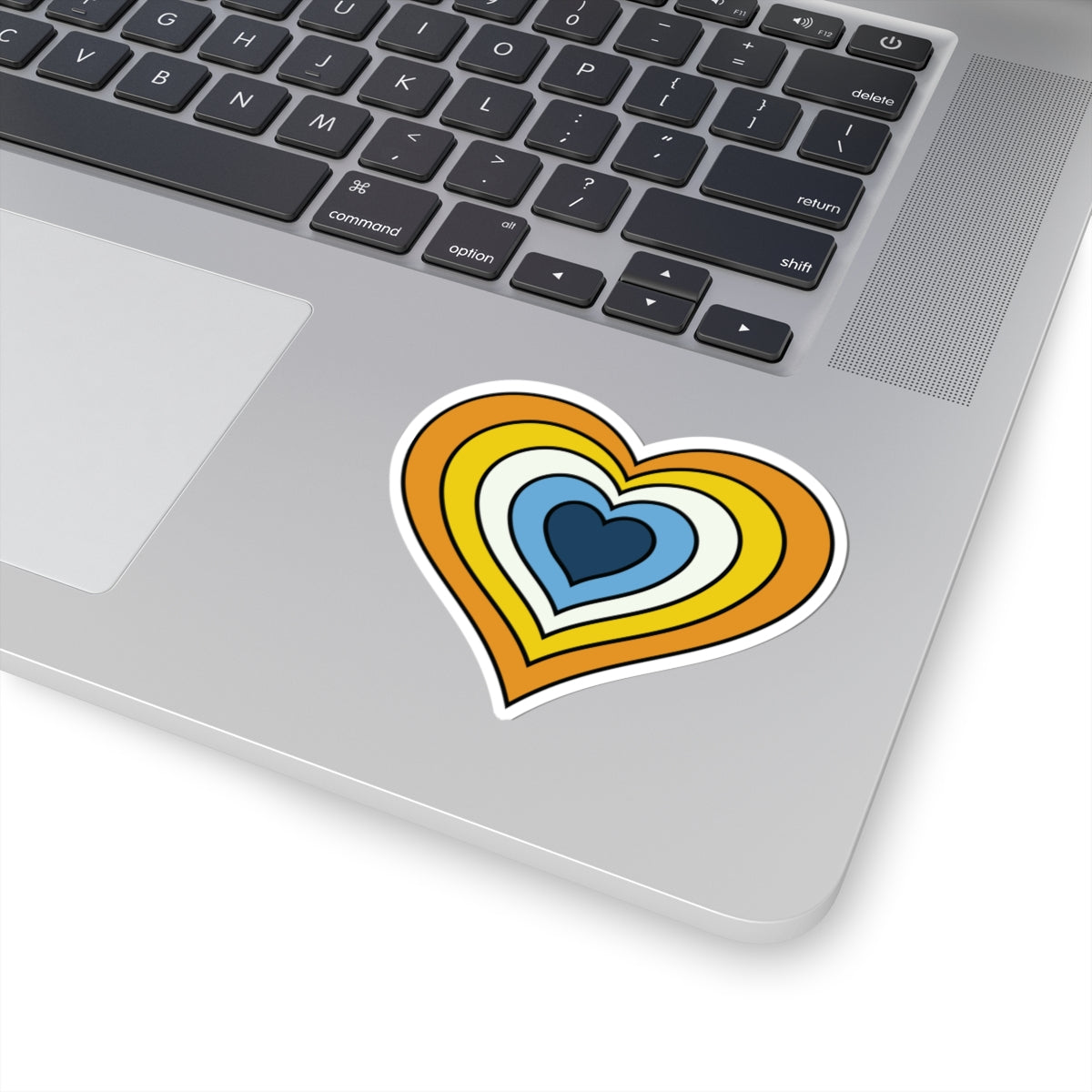 Close-up of the bottom right corner of a silver laptop featuring a 3 inch by 3 inch pride flag heart sticker in an Aroace flag style applied to the surface.