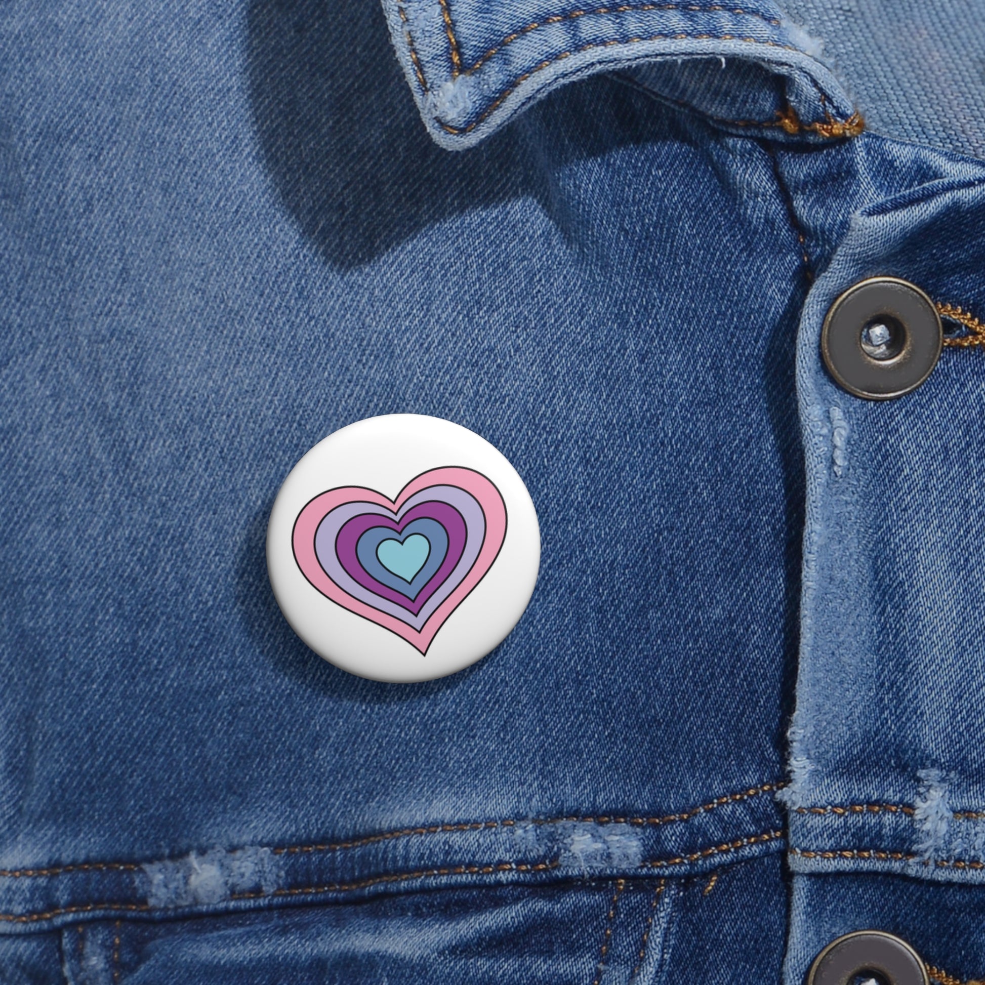 Close-up of a white circular pride button featuring a nested heart design inspired by the Omnigender pride flag in pink, purples and blues, attached to a blue denim jacket at the right chest with the button clearly visible.