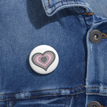 Close-up of a white circular pride button featuring a nested heart design inspired by the Demigirl pride flag in grays, white and pink, attached to a blue denim jacket at the right chest with the button clearly visible.