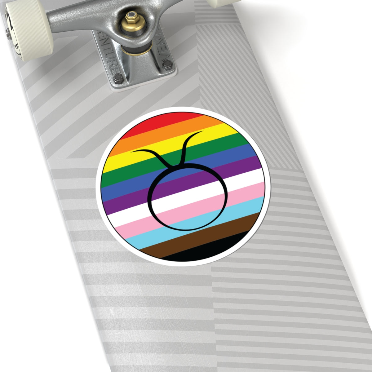 Close-up of the bottom of a silver skateboard featuring a 6 inch by 6 inch circular zodiac sticker with a centered Taurus glyph on a progress pride flag style background applied to the surface.