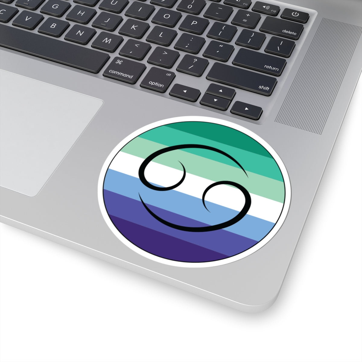 Close-up of the bottom right corner of a silver laptop featuring a 4 inch by 4 inch circular zodiac sticker with a centered Cancer glyph on a Gay Men pride flag style background applied to the surface.