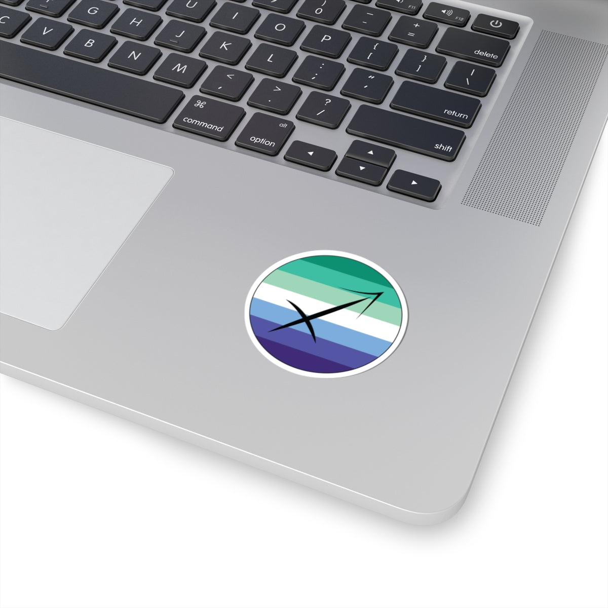 Close-up of the bottom right corner of a silver laptop featuring a 2 inch by 2 inch circular zodiac sticker with a centered Sagittarius glyph on a Gay Men pride flag style background applied to the surface.