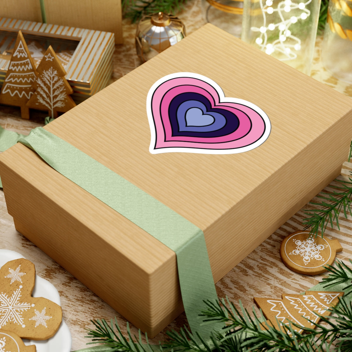 Gift box with a green ribbon on a festive background with cookies and Christmas decorations. Heart-shaped Omnisexual pride sticker centered on the box.