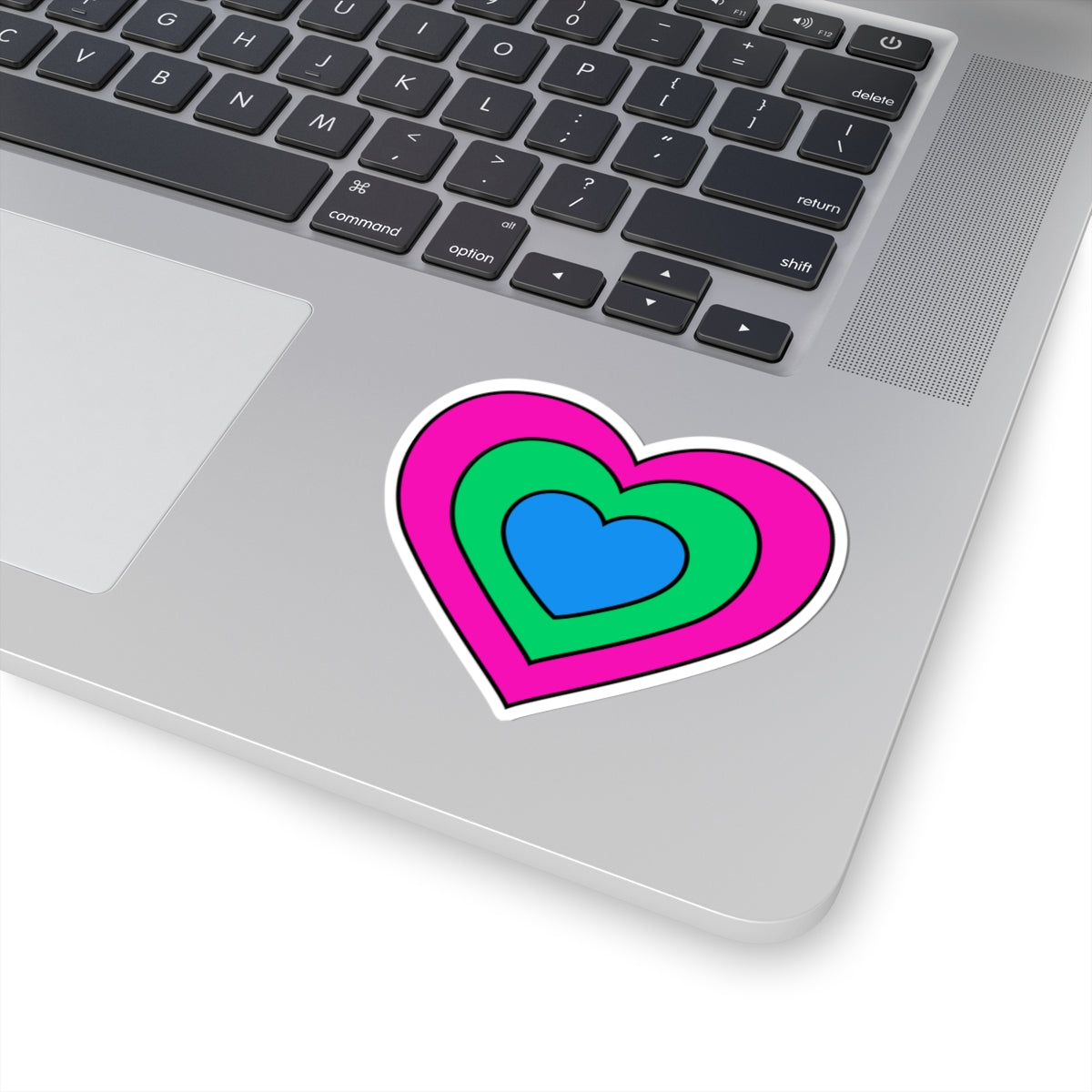Close-up of the bottom right corner of a silver laptop featuring a 3 inch by 3 inch pride flag heart sticker in a Polysexual flag style applied to the surface.