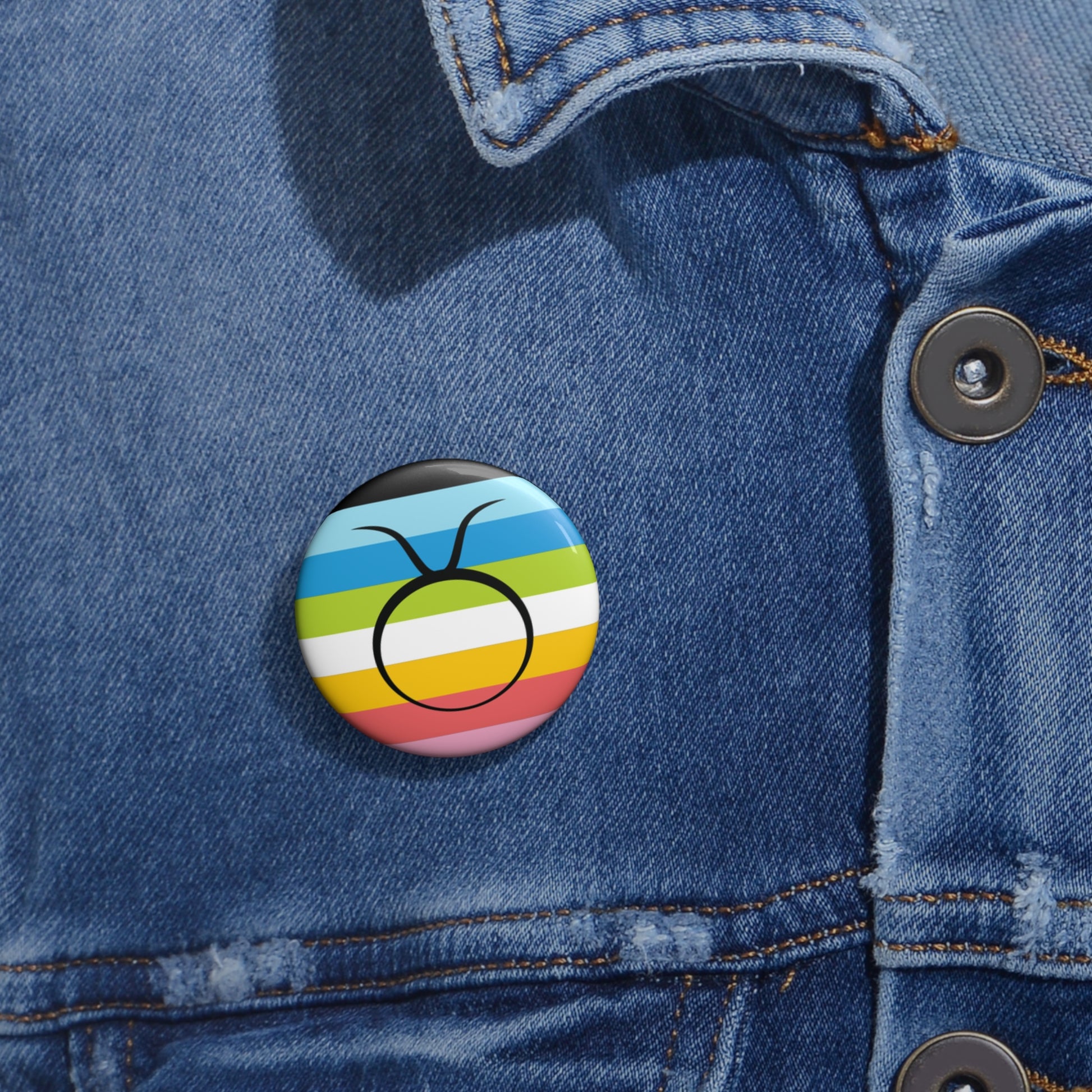 Close-up of a white circular pride button featuring the Taurus zodiac glyph centered over a queer pride flag background in pinks, yellow, white, green, blues, and black, attached to a blue denim jacket at the right chest with the button clearly visible.