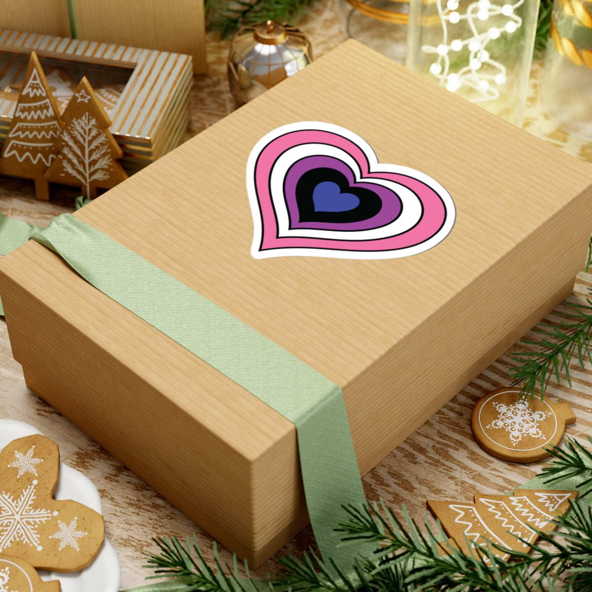 Gift box with a green ribbon on a festive background with cookies and Christmas decorations. Heart-shaped Genderfluid pride sticker centered on the box.