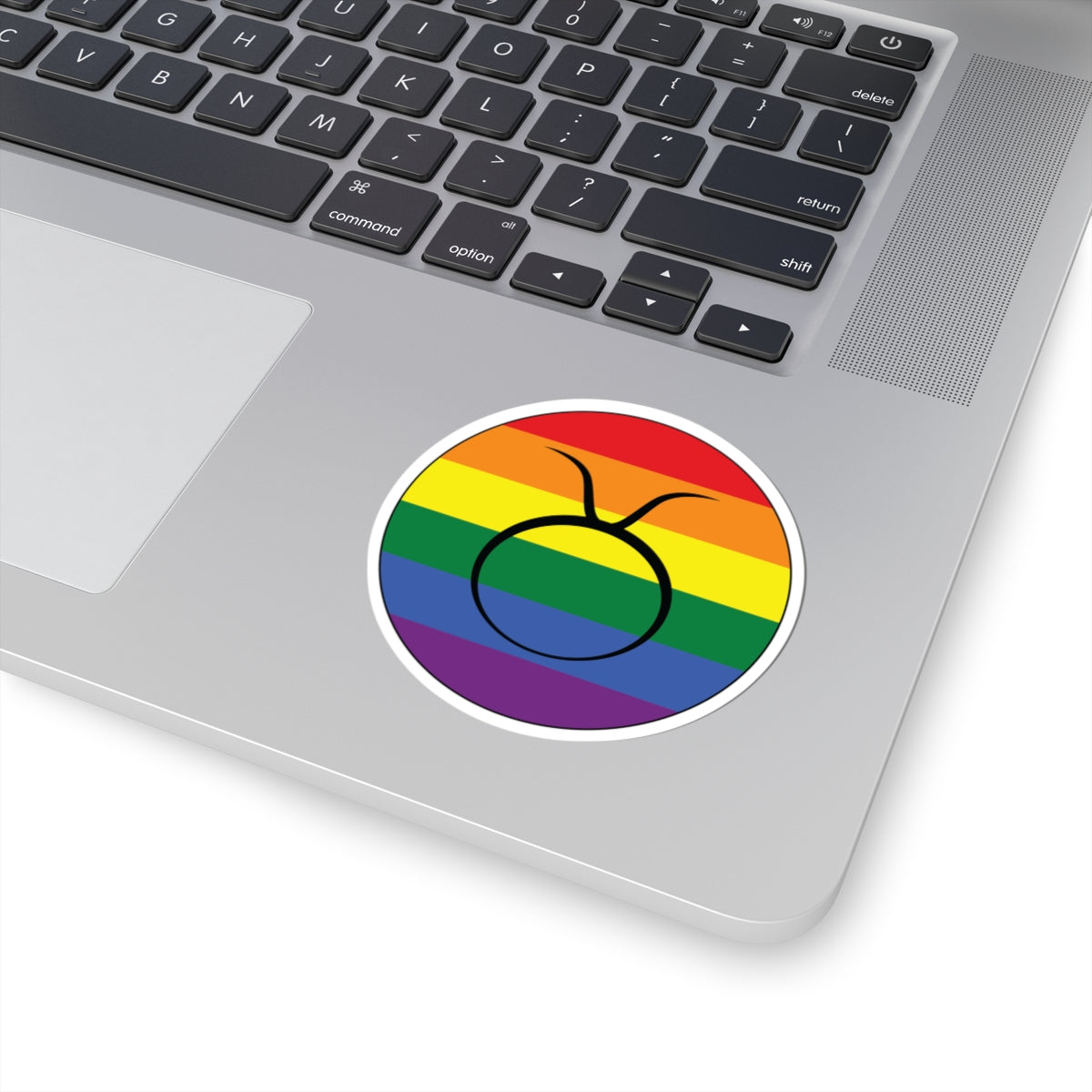 Close-up of the bottom right corner of a silver laptop featuring a 3 inch by 3 inch circular zodiac sticker with a centered Taurus glyph on a Rainbow pride flag style background applied to the surface.
