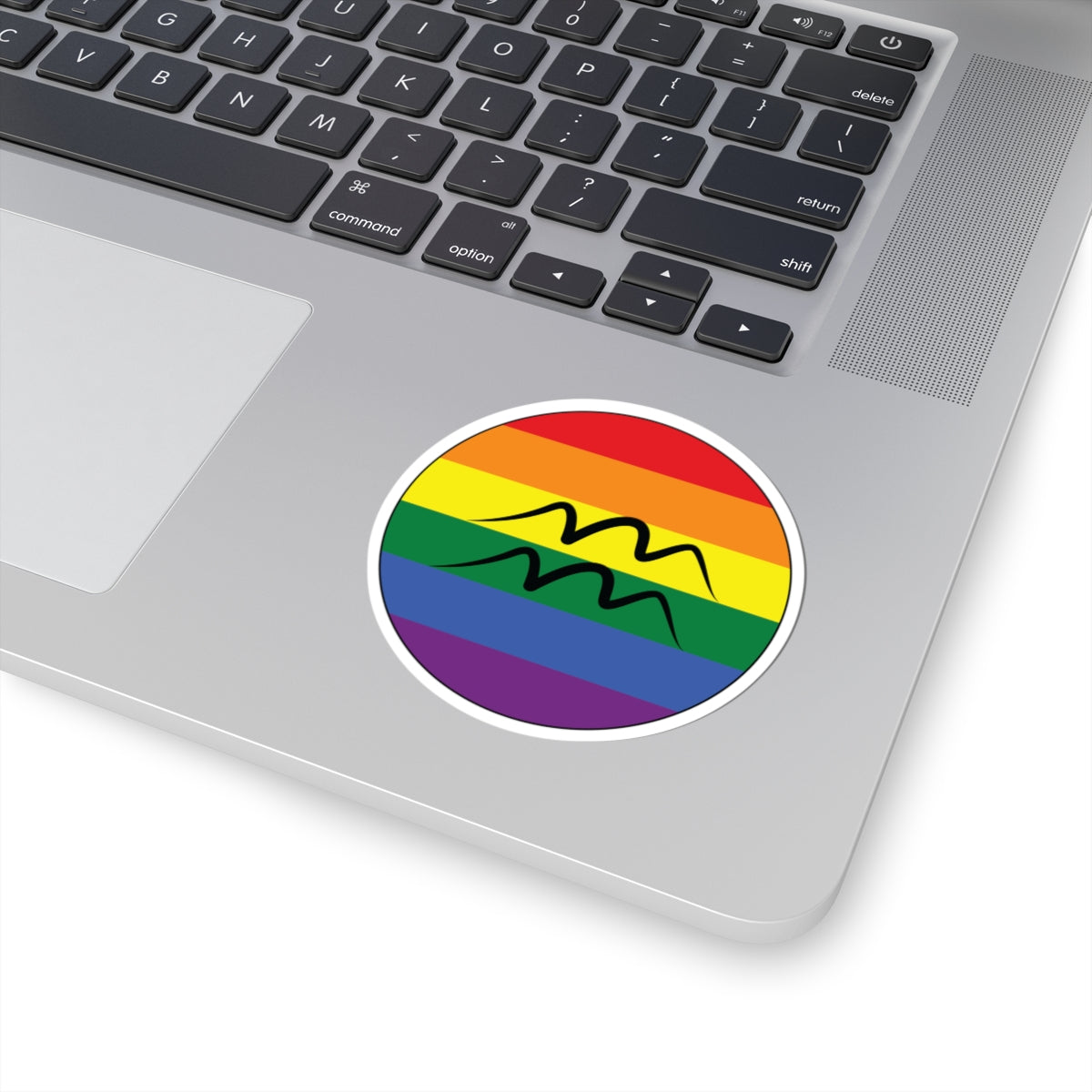 Close-up of the bottom right corner of a silver laptop featuring a 3 inch by 3 inch circular zodiac sticker with a centered Aquarius glyph on a Rainbow pride flag style background applied to the surface.