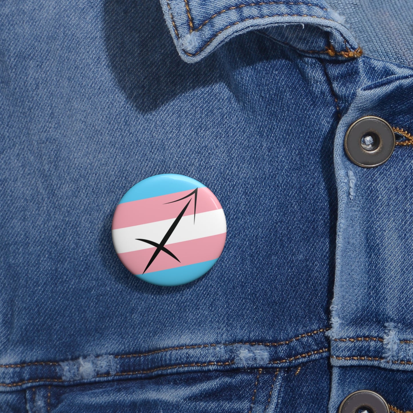 Close-up of a white circular pride button featuring the Sagittarius zodiac glyph centered over a transgender pride flag background in light blue, pink, and white, attached to a blue denim jacket at the right chest with the button clearly visible.