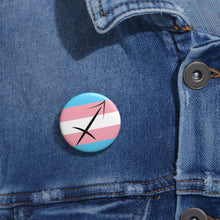 Close-up of a white circular pride button featuring the Sagittarius zodiac glyph centered over a transgender pride flag background in light blue, pink, and white, attached to a blue denim jacket at the right chest with the button clearly visible.