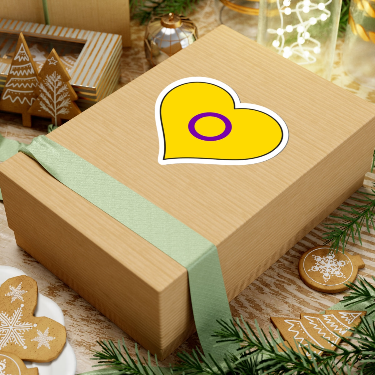 Gift box with a green ribbon on a festive background with cookies and Christmas decorations. Heart-shaped Intersex pride sticker centered on the box.