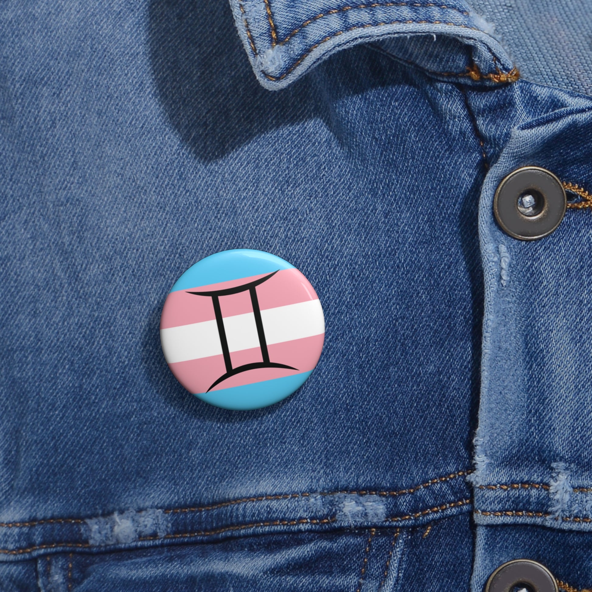 Close-up of a white circular pride button featuring the Gemini zodiac glyph centered over a transgender pride flag background in light blue, pink, and white, attached to a blue denim jacket at the right chest with the button clearly visible.