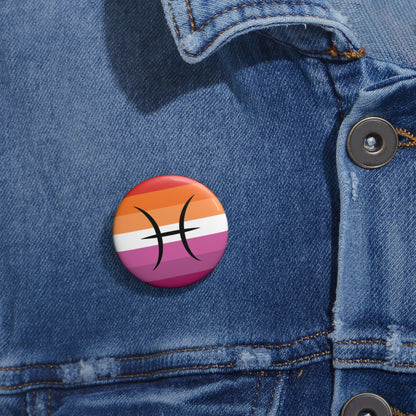 Close-up of a white circular pride button featuring the Pisces zodiac glyph centered over a lesbian pride flag background in oranges, white, and pinks attached to a blue denim jacket at the right chest with the button clearly visible.