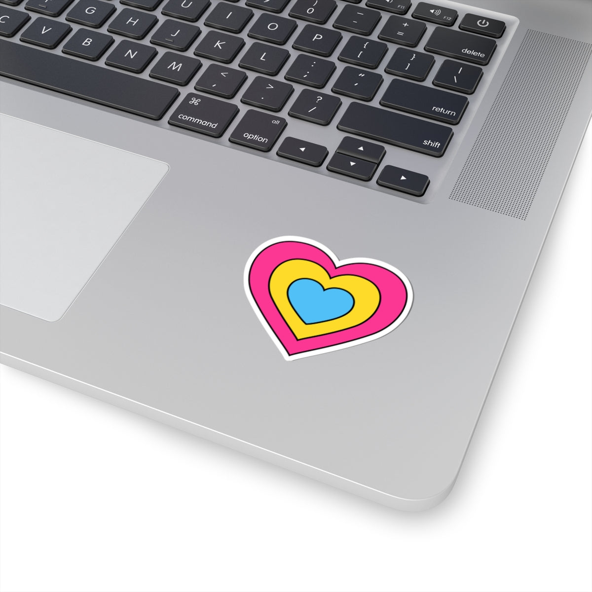 Close-up of the bottom right corner of a silver laptop featuring a 2 inch by 2 inch pride flag heart sticker in a Pansexual flag style applied to the surface.