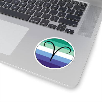 Close-up of the bottom right corner of a silver laptop featuring a 3 inch by 3 inch circular zodiac sticker with a centered Aires glyph on a Gay Men pride flag style background applied to the surface.