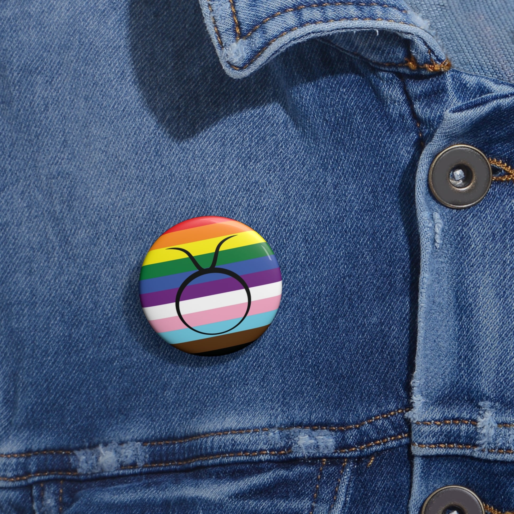 Close-up of a white circular pride button featuring the Taurus zodiac glyph centered over a Progress Pride flag background in rainbow stripes with black, brown, light blue, pink, and white elements, attached to a blue denim jacket at the right chest with the button clearly visible.