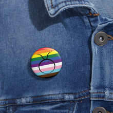 Close-up of a white circular pride button featuring the Taurus zodiac glyph centered over a Progress Pride flag background in rainbow stripes with black, brown, light blue, pink, and white elements, attached to a blue denim jacket at the right chest with the button clearly visible.
