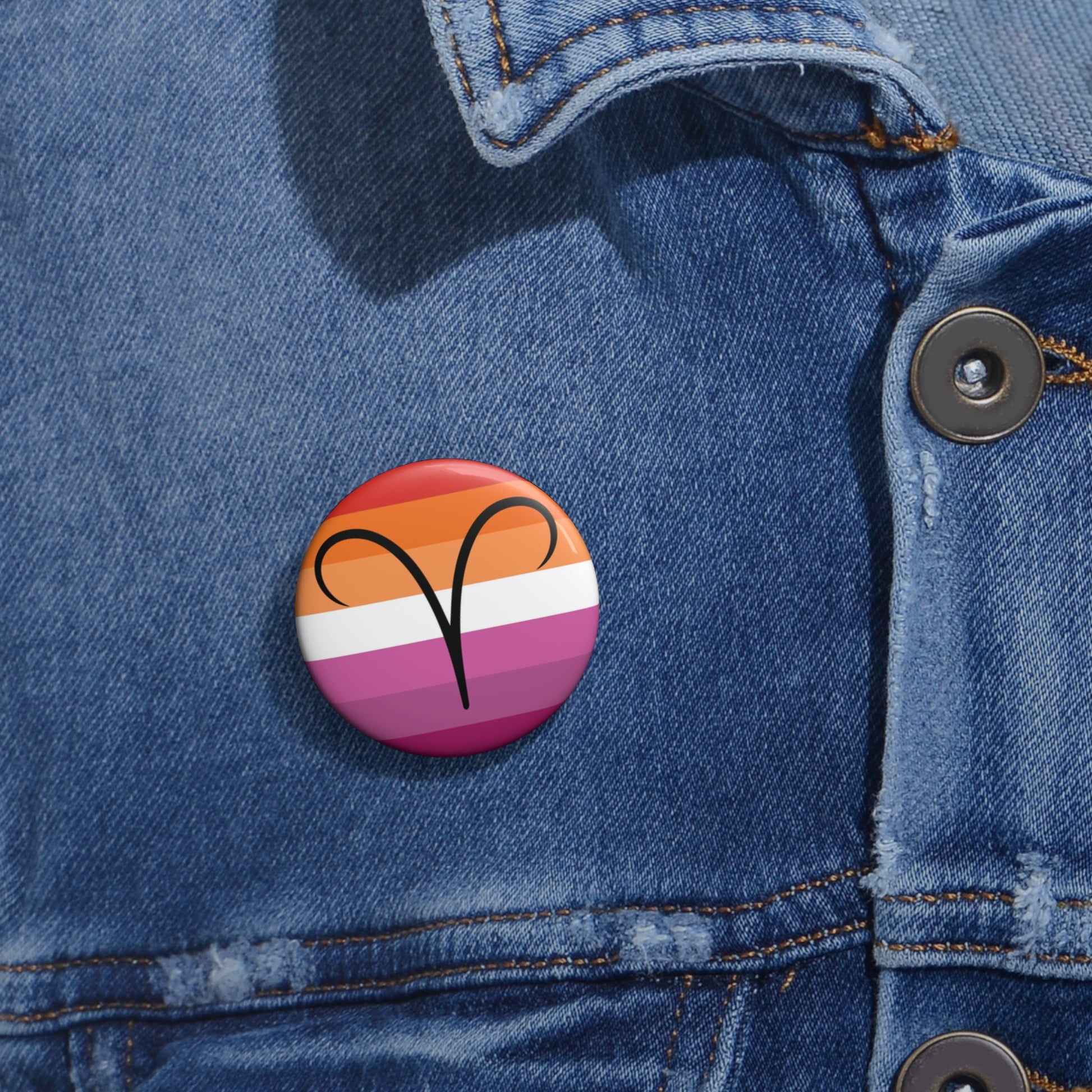 Close-up of a white circular pride button featuring the Aires zodiac glyph centered over a lesbian pride flag background in oranges, white, and pinks attached to a blue denim jacket at the right chest with the button clearly visible.
