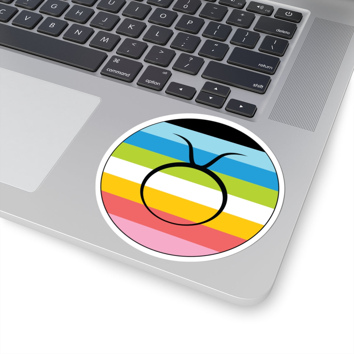 Close-up of the bottom right corner of a silver laptop featuring a 4 inch by 4 inch circular zodiac sticker with a centered Taurus glyph on a Queer pride flag style background applied to the surface.