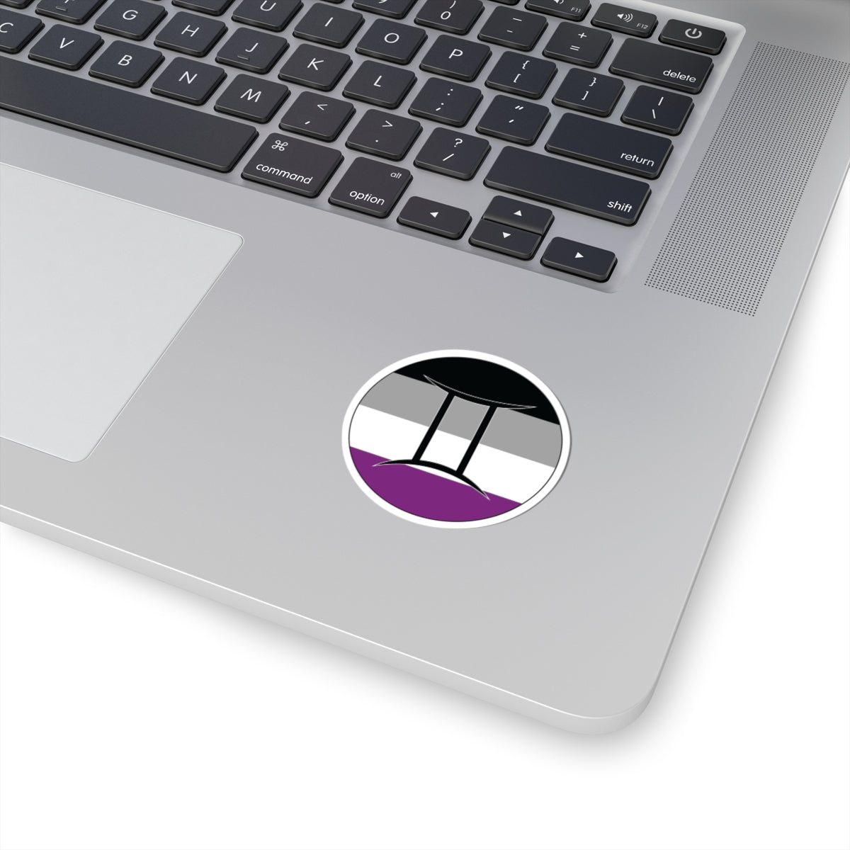 Close-up of the bottom right corner of a silver laptop featuring a 2 inch by 2 inch circular zodiac sticker with a centered Gemini glyph on an Asexual pride flag style background applied to the surface.