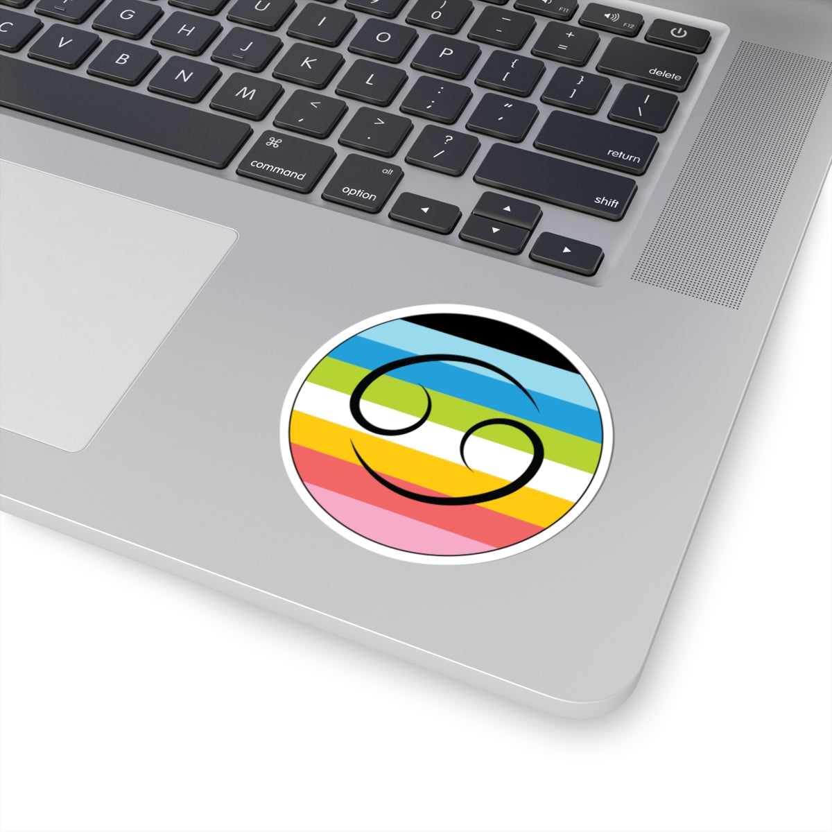 Close-up of the bottom right corner of a silver laptop featuring a 3 inch by 3 inch circular zodiac sticker with a centered Cancer glyph on a Queer pride flag style background applied to the surface.