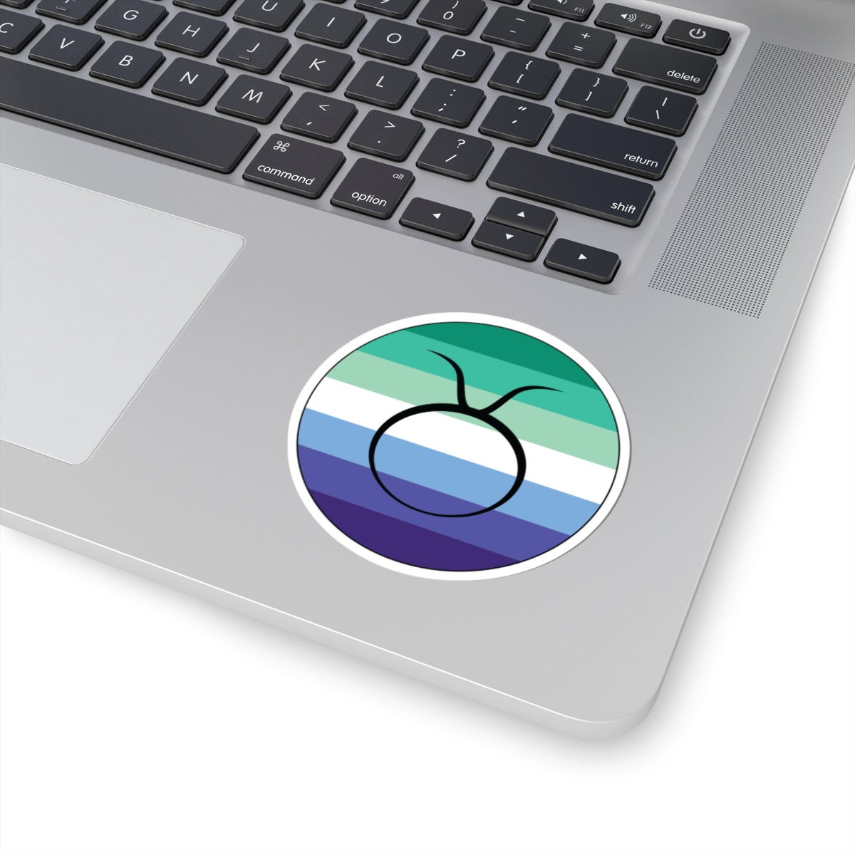Close-up of the bottom right corner of a silver laptop featuring a 3 inch by 3 inch circular zodiac sticker with a centered Taurus glyph on a Gay Men pride flag style background applied to the surface.