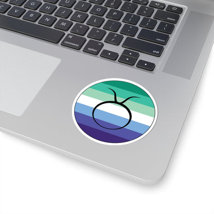 Close-up of the bottom right corner of a silver laptop featuring a 3 inch by 3 inch circular zodiac sticker with a centered Taurus glyph on a Gay Men pride flag style background applied to the surface.