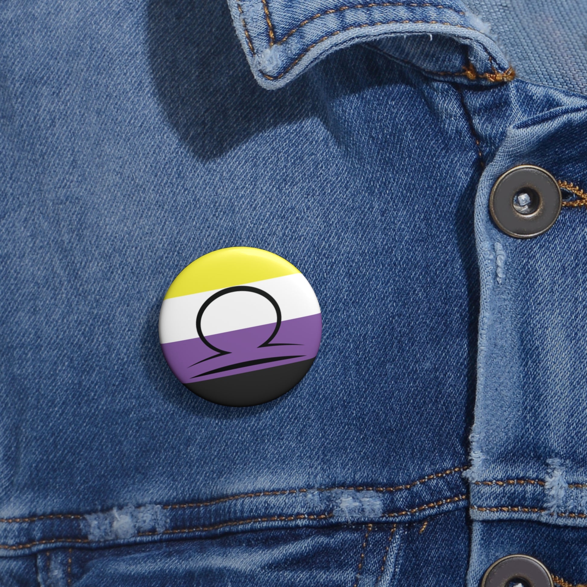 Close-up of a white circular pride button featuring the Libra zodiac glyph centered over a nonbinary pride flag background in yellow, white, purple, and black, attached to a blue denim jacket at the right chest with the button clearly visible.