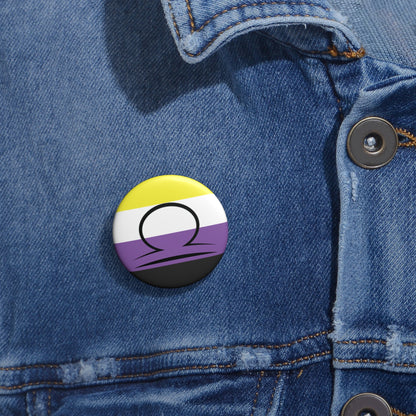 Close-up of a white circular pride button featuring the Libra zodiac glyph centered over a nonbinary pride flag background in yellow, white, purple, and black, attached to a blue denim jacket at the right chest with the button clearly visible.