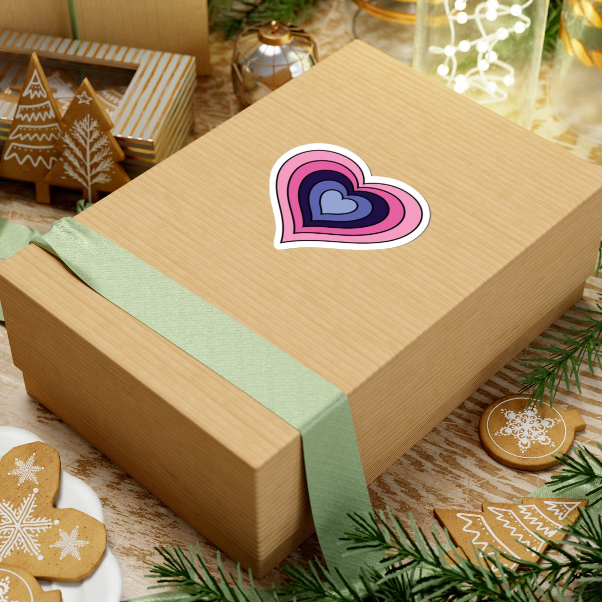 Gift box with a green ribbon on a festive background with cookies and Christmas decorations. Heart-shaped Omnisexual pride sticker centered on the box.