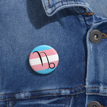Close-up of a white circular pride button featuring the Capricorn zodiac glyph centered over a transgender pride flag background in light blue, pink, and white, attached to a blue denim jacket at the right chest with the button clearly visible.