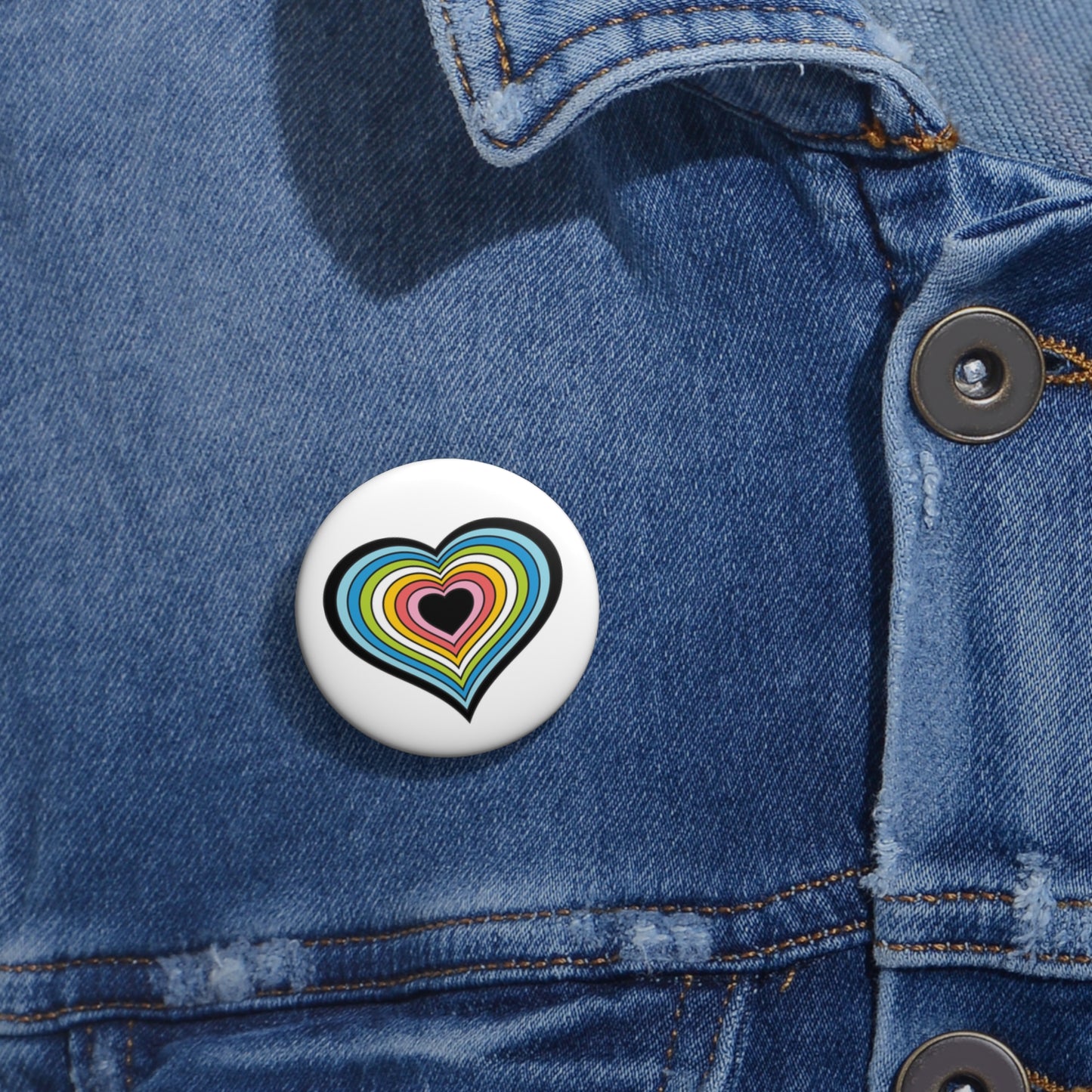 Close-up of a white circular pride button featuring a nested heart design inspired by the Queer pride flag in black, blues, lime green, white, and pinks, attached to a blue denim jacket at the right chest with the button clearly visible.