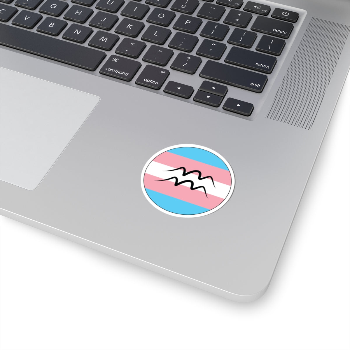 Close-up of the bottom right corner of a silver laptop featuring a 2 inch by 2 inch circular zodiac sticker with a centered Aquarius glyph on a Transgender pride flag style background applied to the surface.
