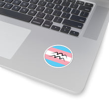 Close-up of the bottom right corner of a silver laptop featuring a 2 inch by 2 inch circular zodiac sticker with a centered Aquarius glyph on a Transgender pride flag style background applied to the surface.