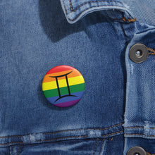 Close-up of a white circular pride button featuring the Gemini zodiac glyph centered over a rainbow pride flag background in red, orange, yellow, green, blue, and purple, attached to a blue denim jacket at the right chest with the button clearly visible.