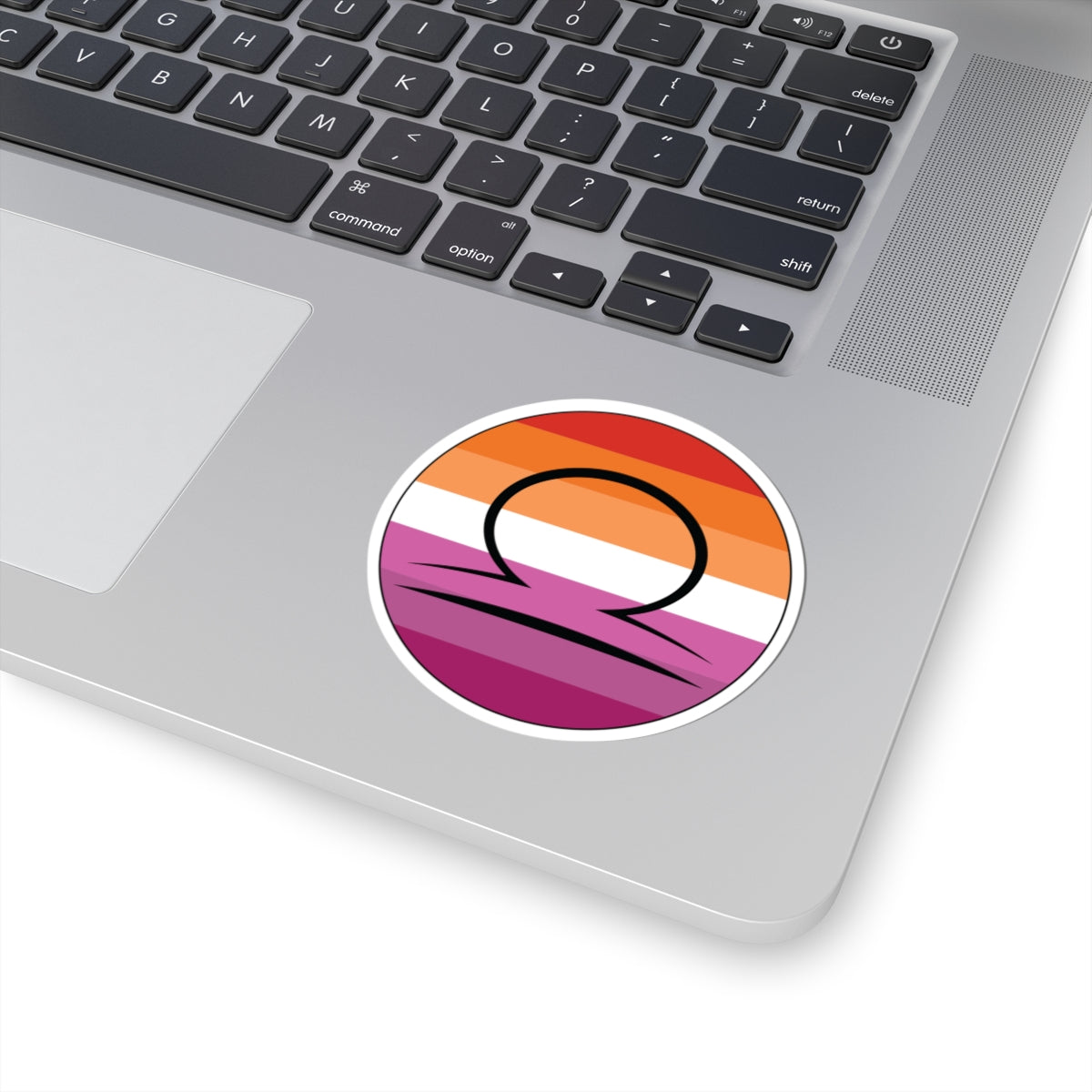Close-up of the bottom right corner of a silver laptop featuring a 3 inch by 3 inch circular zodiac sticker with a centered Libra glyph on a Lesbian pride flag style background applied to the surface.