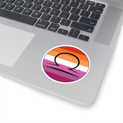 Close-up of the bottom right corner of a silver laptop featuring a 3 inch by 3 inch circular zodiac sticker with a centered Libra glyph on a Lesbian pride flag style background applied to the surface.