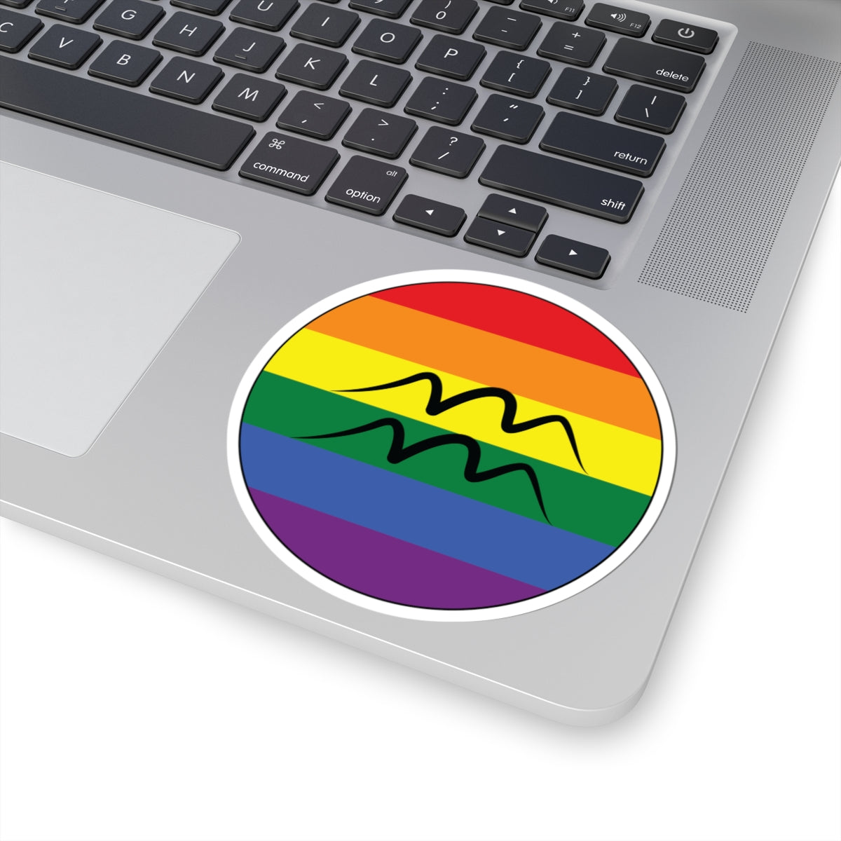 Close-up of the bottom right corner of a silver laptop featuring a 4 inch by 4 inch circular zodiac sticker with a centered Aquarius glyph on a Rainbow pride flag style background applied to the surface.