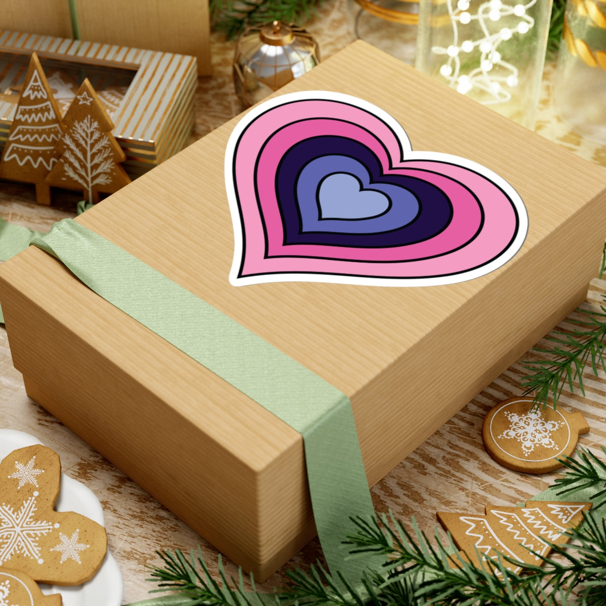 Gift box with a green ribbon on a festive background with cookies and Christmas decorations. Heart-shaped Omnisexual pride sticker centered on the box.