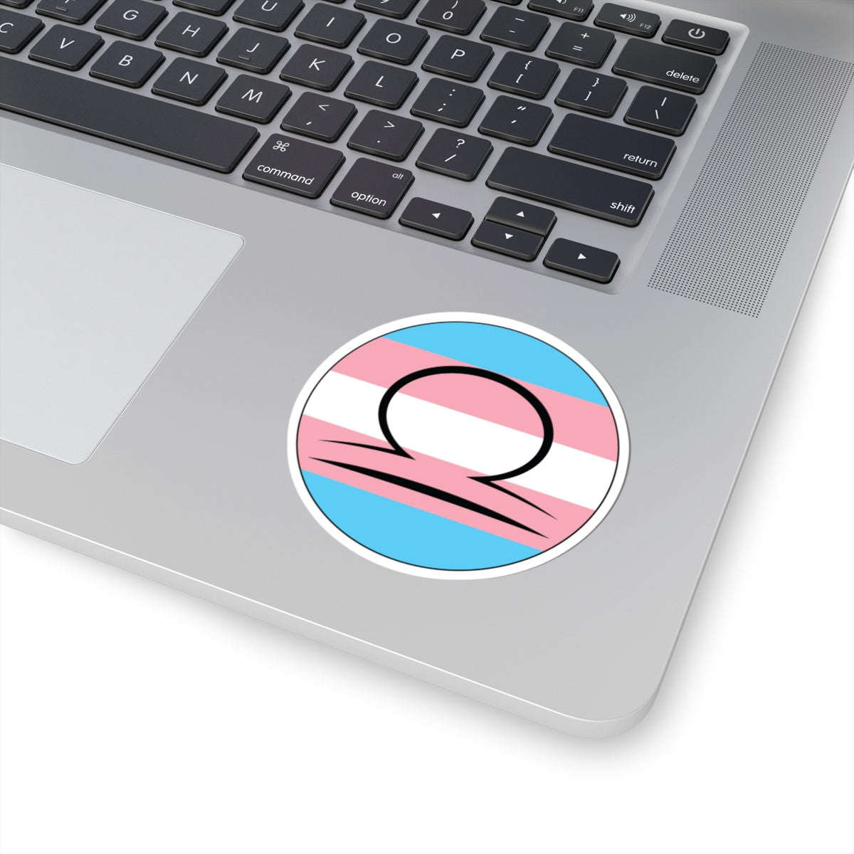 Close-up of the bottom right corner of a silver laptop featuring a 3 inch by 3 inch circular zodiac sticker with a centered Libra glyph on a Transgender pride flag style background applied to the surface.