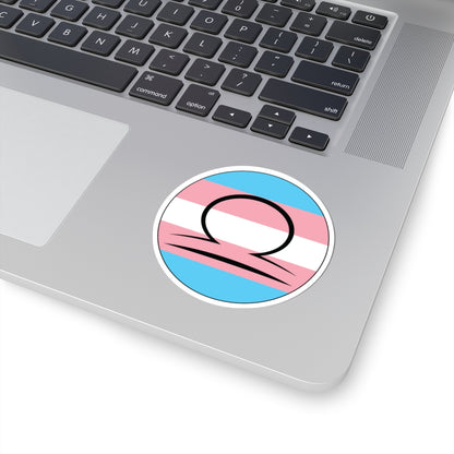 Close-up of the bottom right corner of a silver laptop featuring a 3 inch by 3 inch circular zodiac sticker with a centered Libra glyph on a Transgender pride flag style background applied to the surface.