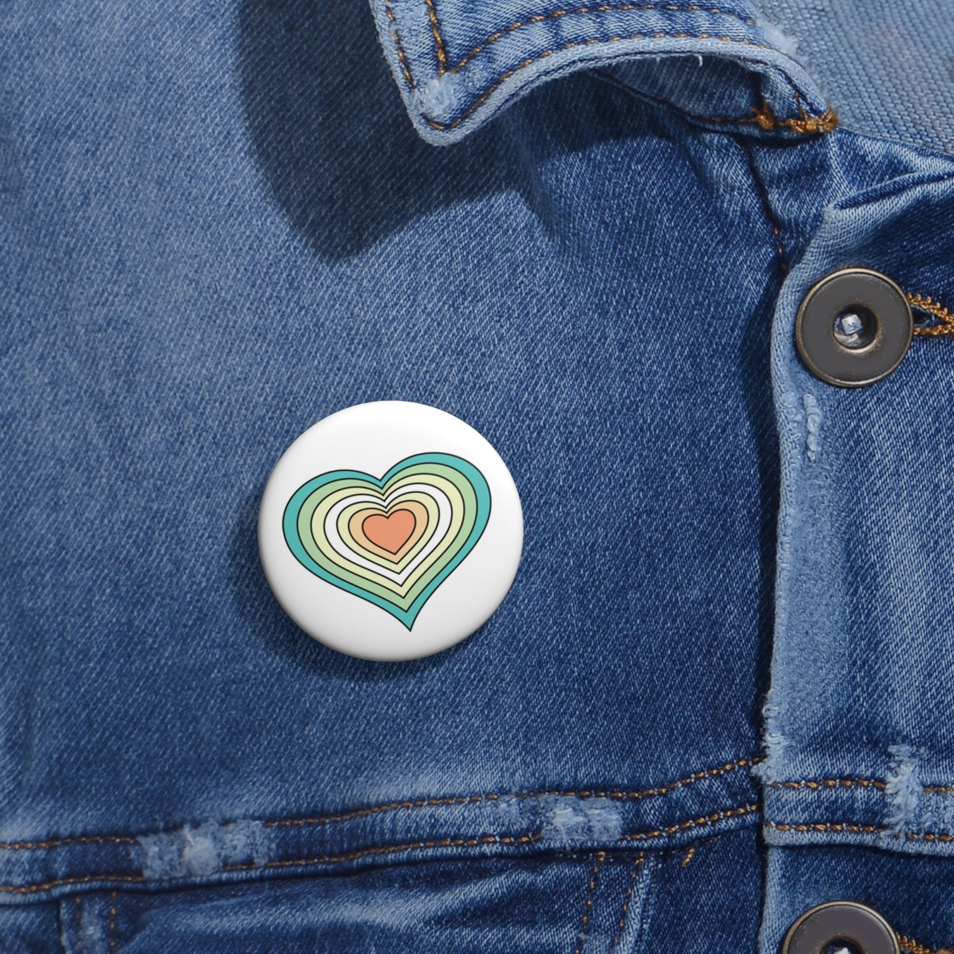 Close-up of a white circular pride button featuring a nested heart design inspired by the Genderfloren pride flag in greens, white, yellow and orange, attached to a blue denim jacket at the right chest with the button clearly visible.