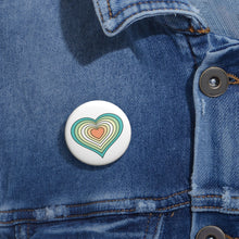 Close-up of a white circular pride button featuring a nested heart design inspired by the Genderfloren pride flag in greens, white, yellow and orange, attached to a blue denim jacket at the right chest with the button clearly visible.