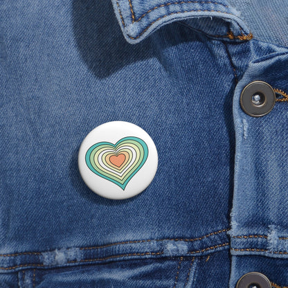Close-up of a white circular pride button featuring a nested heart design inspired by the Genderfloren pride flag in greens, white, yellow and orange, attached to a blue denim jacket at the right chest with the button clearly visible.