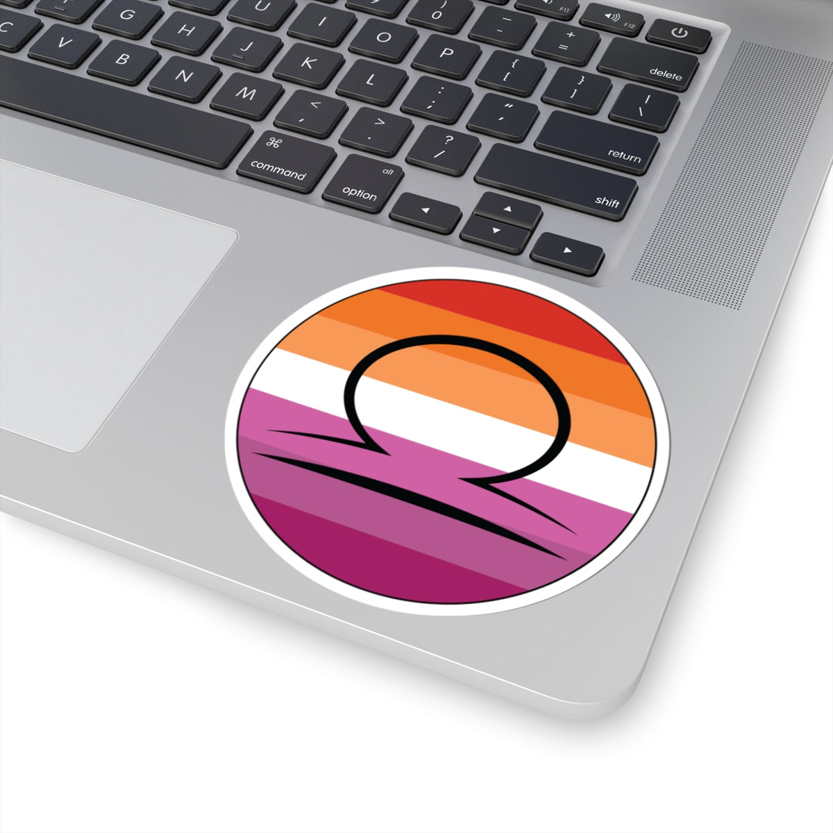 Close-up of the bottom right corner of a silver laptop featuring a 4 inch by 4 inch circular zodiac sticker with a centered Libra glyph on a Lesbian pride flag style background applied to the surface.
