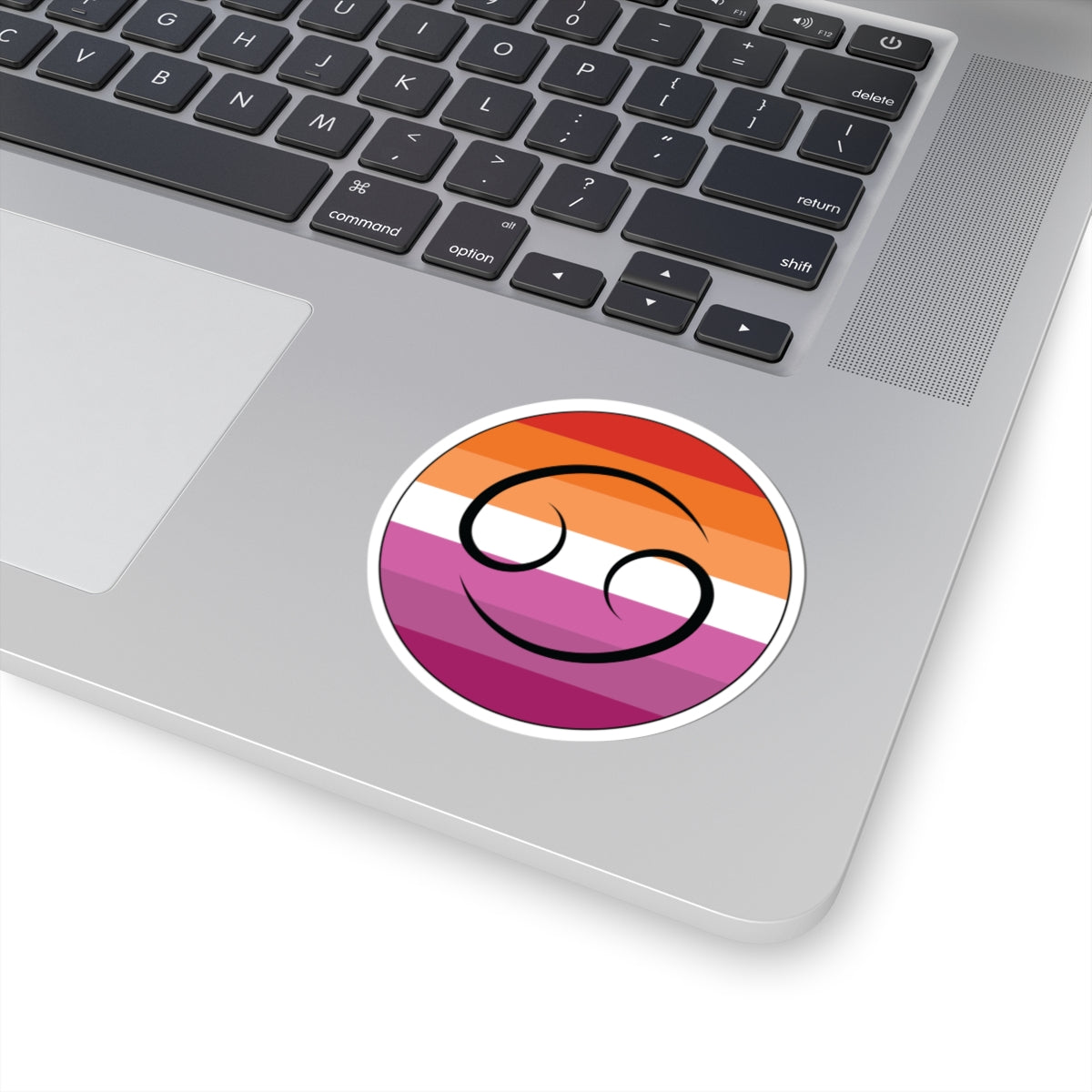Close-up of the bottom right corner of a silver laptop featuring a 3 inch by 3 inch circular zodiac sticker with a centered Cancer glyph on a Lesbian pride flag style background applied to the surface.