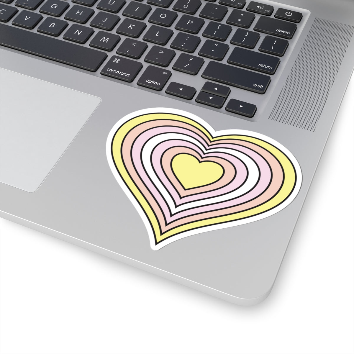 Close-up of the bottom right corner of a silver laptop featuring a 4 inch by 4 inch pride flag heart sticker in a Pangender flag style applied to the surface.
