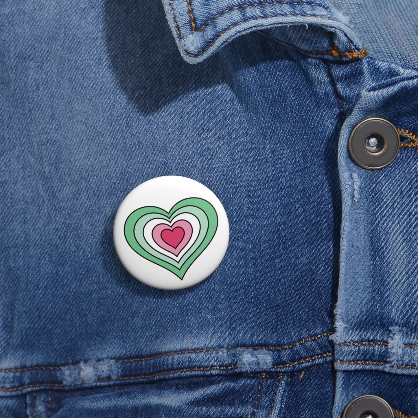 Close-up of a white circular pride button featuring a nested heart design inspired by the Abrosexual pride flag in greens, white, and pinks, attached to a blue denim jacket at the right chest with the button clearly visible.
