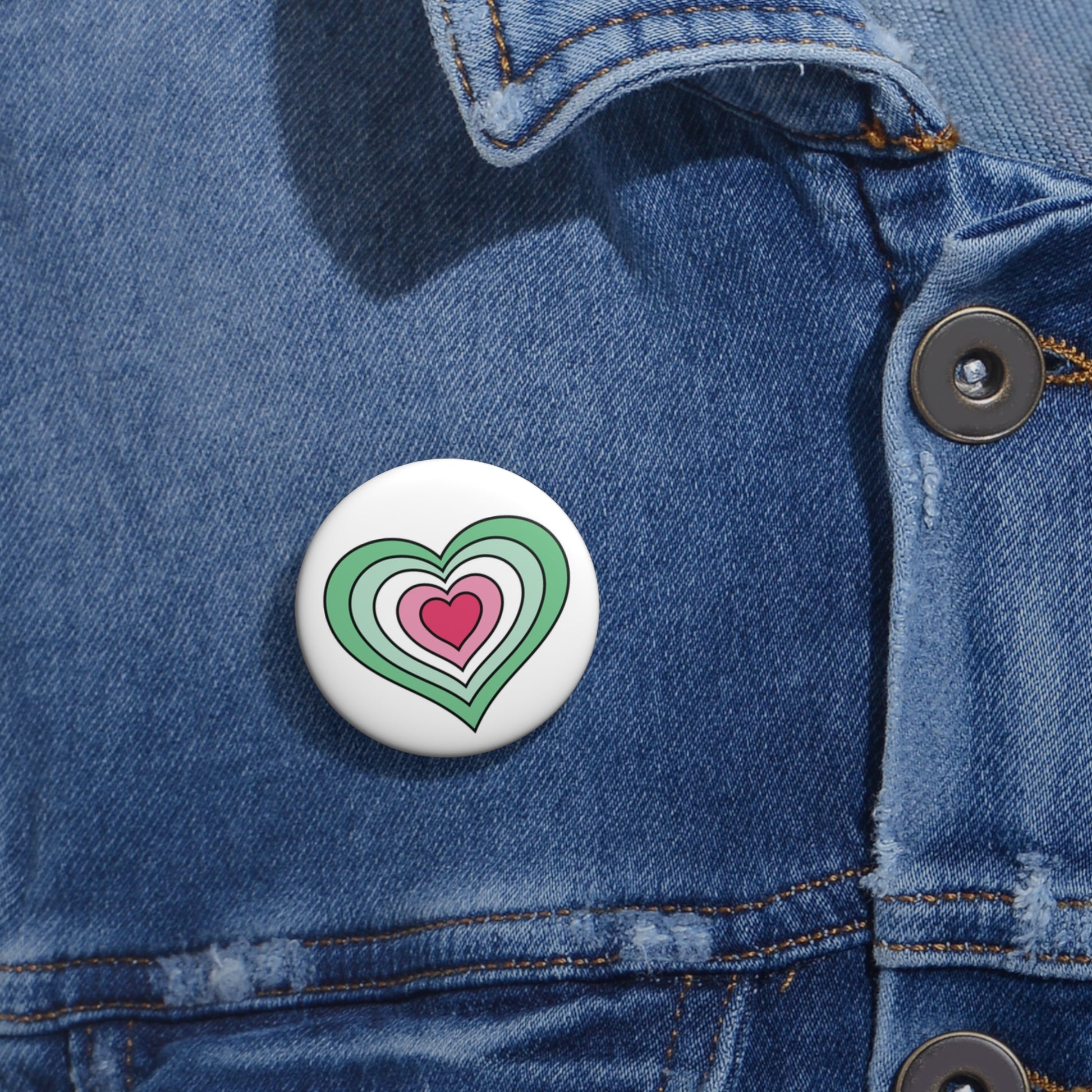 Close-up of a white circular pride button featuring a nested heart design inspired by the Abrosexual pride flag in greens, white, and pinks, attached to a blue denim jacket at the right chest with the button clearly visible.